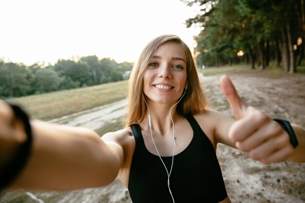 Bella ragazza sorridente in abiti sportivi che prendono un selfie, mostrante un dito grosso