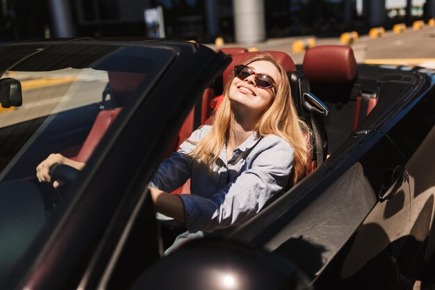 Bella ragazza sorridente in occhiali da sole che guarda felicemente a porte chiuse mentre guida un'auto cabriolet per le strade della città all'aperto