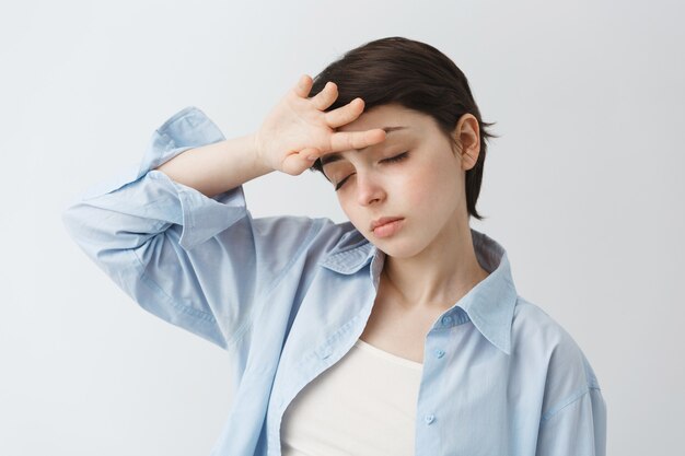 Bella ragazza stanca con i capelli corti chiude gli occhi e si tocca la fronte, ha febbre o mal di testa, sintomi covid-19