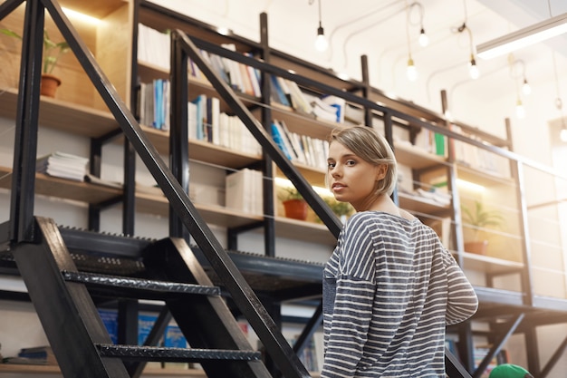 Bella ragazza studentessa bionda con i capelli corti in camicia a strisce casual trascorrere del tempo in biblioteca moderna dopo l'università, preparando per gli esami con gli amici. Ragazza che sta vicino alle scale che vanno a prendere