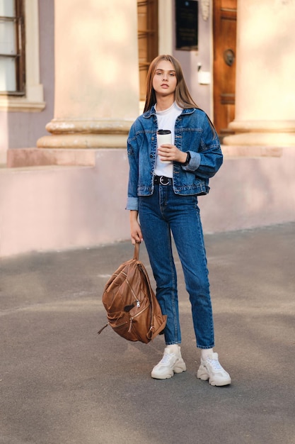 Bella ragazza studentessa elegante e casual in giacca di jeans con zaino che guarda con sicurezza a porte chiuse nell'università all'aperto