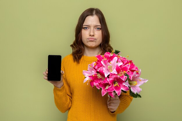 Bella ragazza triste il giorno delle donne felici che tiene il mazzo con il telefono isolato sulla parete verde oliva