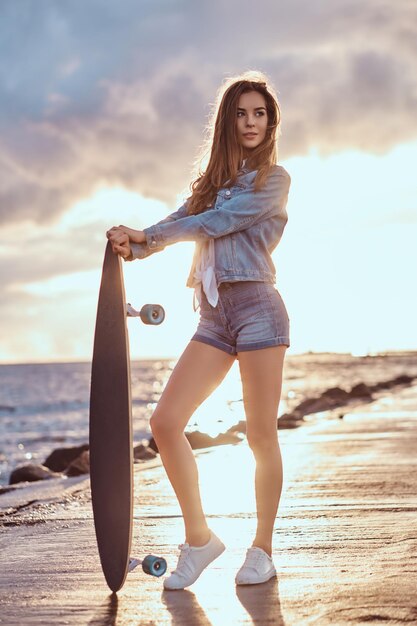 Bella ragazza vestita in pantaloncini di jeans e giacca in posa con uno skateboard sulla spiaggia con tempo nuvoloso durante il tramonto luminoso.