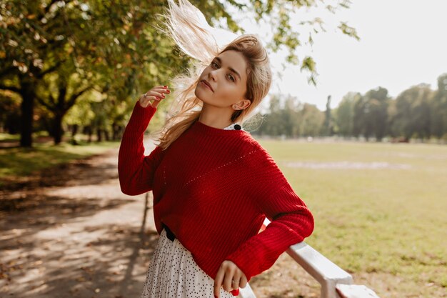 Bella signora che gioca con i suoi capelli all'aperto. Giovane donna bionda in posa volentieri sotto gli alberi.