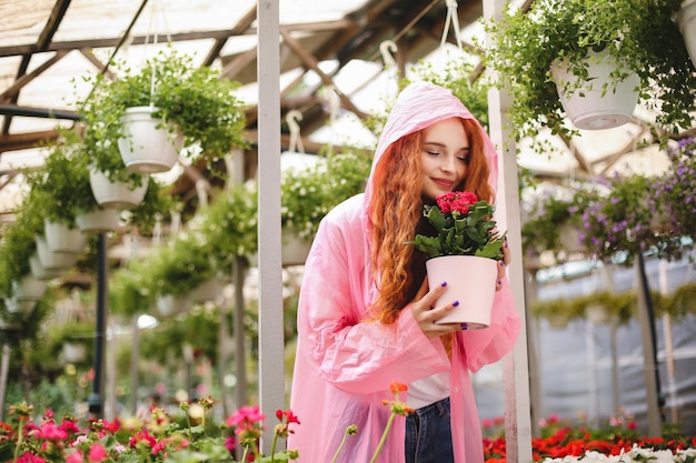 Bella signora con capelli ricci rossi in piedi in impermeabile rosa e fiori in vaso dall'odore sognante mentre trascorre il tempo in serra