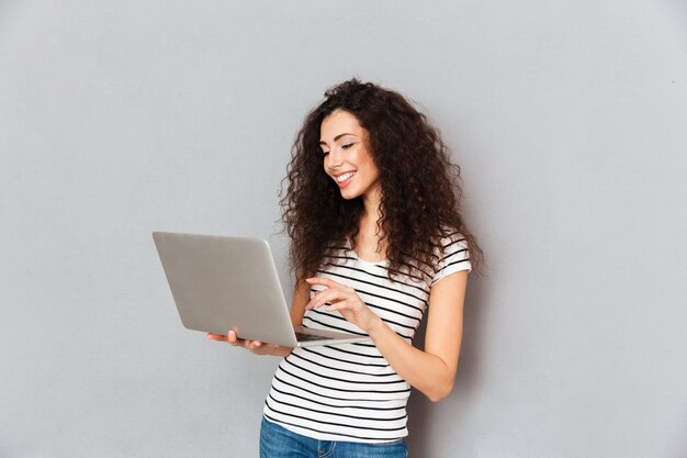 Bella signora con i capelli ricci e-mail con la sua amica utilizzando il computer portatile d'argento essendo isolato su muro grigio