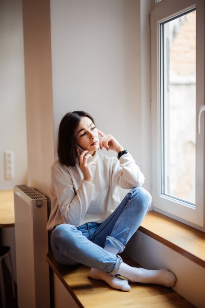 Bella signora dai capelli corti in blue jeans che si siedono sul davanzale e che scrivono messaggio sullo smartphone a casa