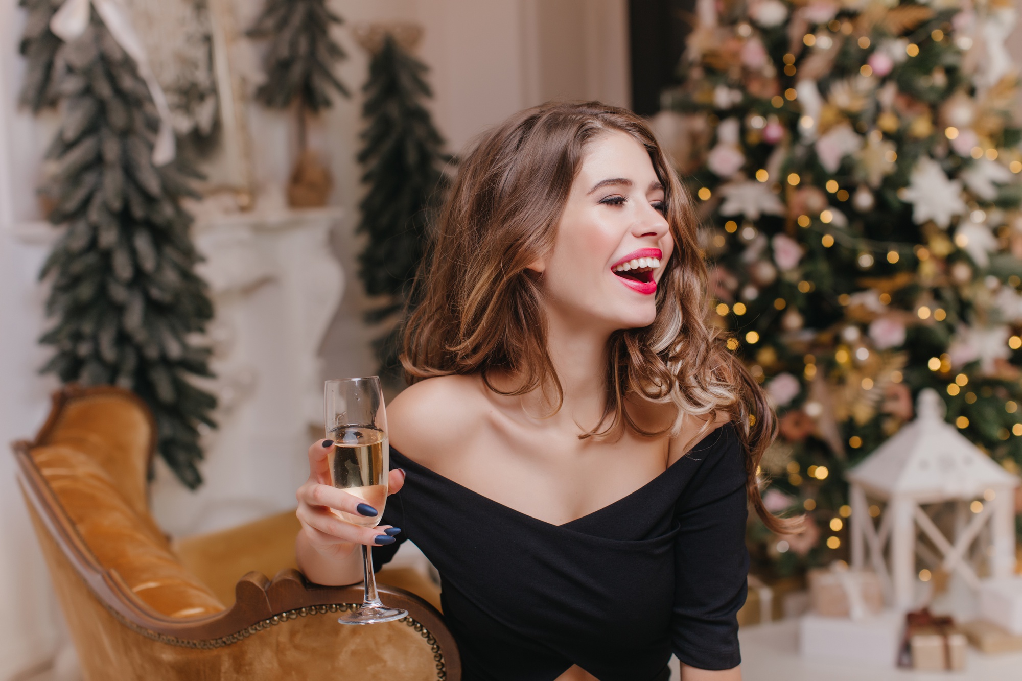 Bella signora dai capelli scuri in abito nero in posa nel giorno di Capodanno con un bicchiere di champagne. Foto dell'interno del modello femminile europeo di bell'aspetto che celebra il Natale a casa e ride.