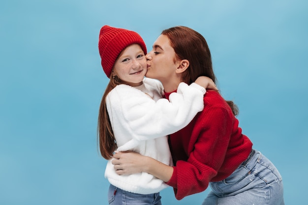 Bella signora elegante in maglione rosso brillante e gonna di jeans che bacia sulla guancia giovane ragazza zenzero con cappello di lana e maglione bianco oversize sulla parete blu