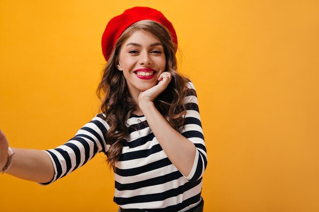 Bella signora in berretto rosso sorridente e prendendo selfie. Bella giovane donna con labbra luminose in cappello alla moda e camicetta a righe in posa.