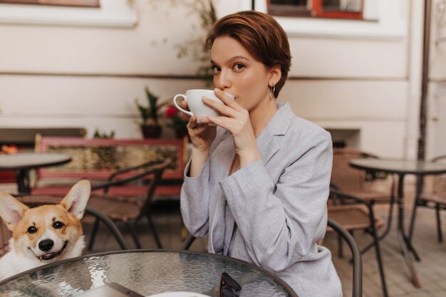 Bella signora in giacca grigia beve caffè in street cafe. La giovane donna in vestito alla moda gode del tè e posa con il corgi all'esterno