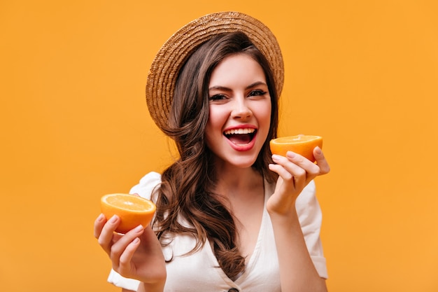 Bella signora in maglietta bianca e cappello di paglia guarda nella telecamera con un sorriso e tiene le fette d'arancia.