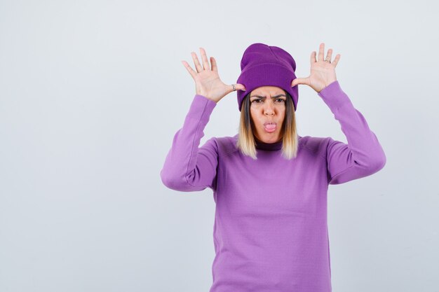 Bella signora in maglione, berretto con le mani vicino alla testa come orecchie, che tira fuori la lingua e sembra scontroso, vista frontale.