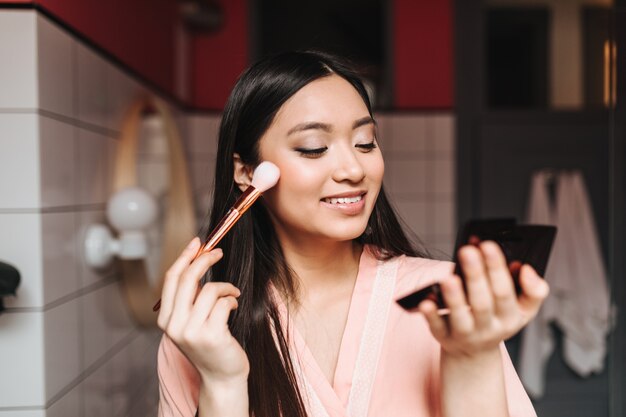 Bella signora in veste di seta rosa con il sorriso fa il trucco in bagno