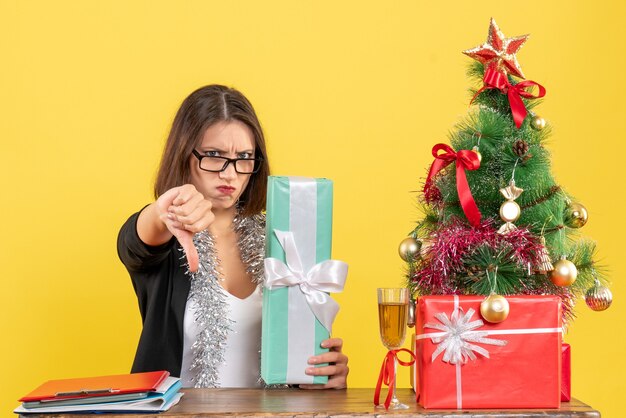 Bella signora in vestito con gli occhiali che mostra il suo regalo facendo un gesto negativo e seduto a un tavolo con un albero di Natale su di esso in ufficio