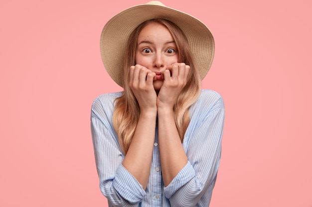 Bella signora nevrotica in cappello e camicia, si morde le unghie, fissa con paura, si sente in ansia per qualcosa, isolata sopra il muro rosa. La giovane donna elegante osserva nervosamente. Persone e sentimenti