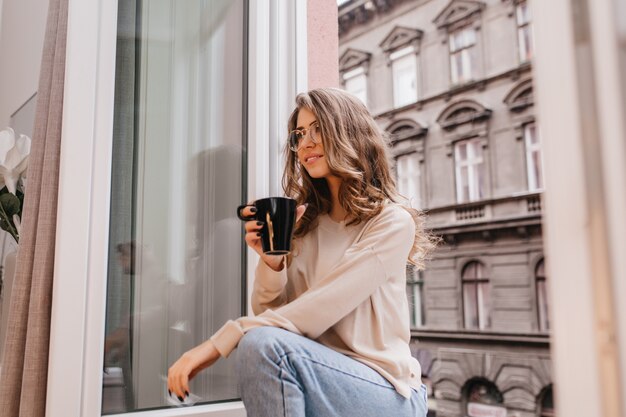 Bella signora sognante in blue jeans che guarda lontano, seduto accanto alla finestra