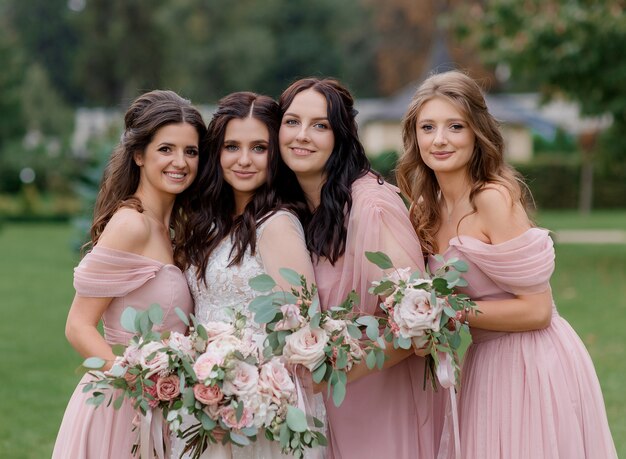 Bella sposa con damigelle vestite in abiti rosa stanno tenendo mazzi di fiori rosa pallido fatti di rose all'aperto