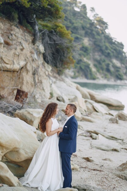 bella sposa dai capelli lunghi in abito bianco con il marito sulla spiaggia vicino a grandi pietre