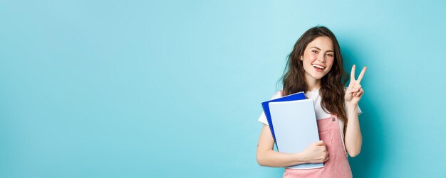Bella studentessa che mostra vsign e quaderni felici sorridenti della tenuta con materiale di studio atten