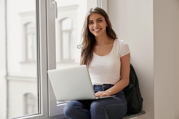 Bella studentessa seduta con il computer portatile sul davanzale