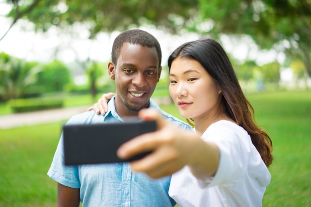 Belle coppie interrazziali sorridenti che prendono selfie nel parco di estate.