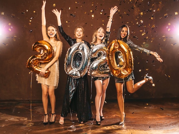 Belle donne che celebrano il nuovo anno. Ragazze bellissime felici in eleganti abiti da festa sexy con palloncini oro e argento 2020, divertendosi alla festa di Capodanno. Festeggiamenti festivi. Mani alzanti