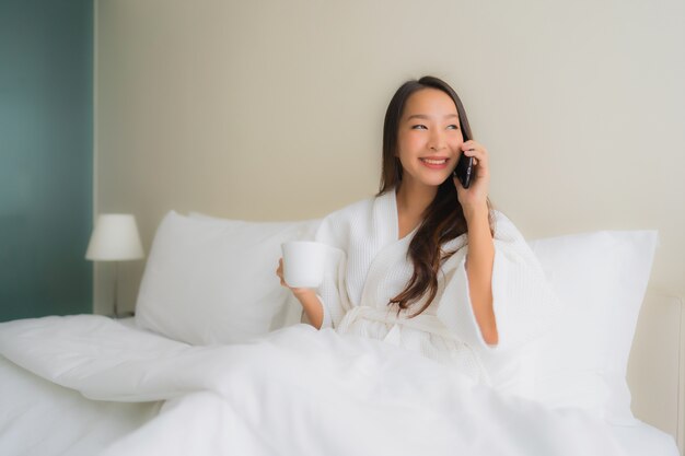 Belle giovani donne asiatiche del ritratto con la tazza e il telefono cellulare di caffè sul letto