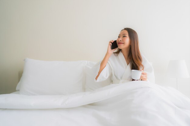 Belle giovani donne asiatiche del ritratto con la tazza e il telefono cellulare di caffè sul letto