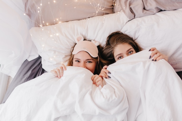 Belle ragazze caucasiche che si nascondono sotto la coltre bianca. Ritratto dell'interno di sorelle giovani sorprendenti in posa mattina.