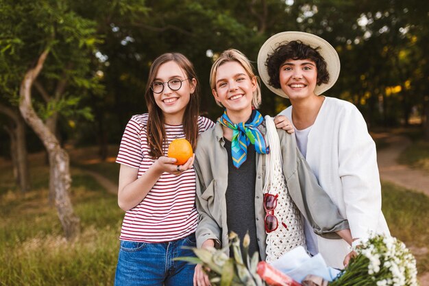 Belle ragazze sorridenti con bicicletta e arancia in mano guardando felicemente nella fotocamera trascorrendo del tempo nel parco