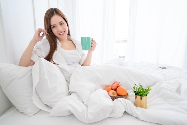 Bellezza asiatica femmina vestito bianco capelli lunghi mano tenere bevanda calda mattina sul letto bianco con vongole e allegro sfondo bianco camera da letto interna