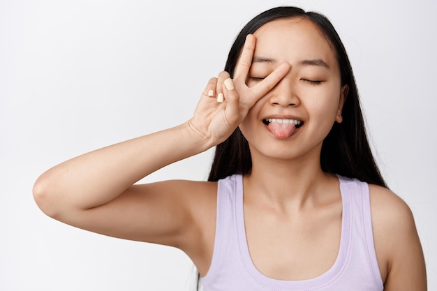 Bellezza attraente viso donna asiatica che mostra il segno di pace e attaccare la lingua sciocco occhi chiusi La ragazza ha una pelle pulita e luminosa senza acne o trucco sfondo bianco