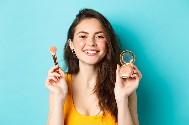 Bellezza. Bellissimo modello femminile con faccia felice, sorridente e mostrando il pennello per il trucco con arrossamenti, in piedi su sfondo blu