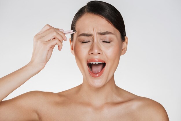 Bellezza della giovane donna stressata con i capelli castani che grida nel dolore mentre pizzicando le sopracciglia usando una pinzetta, isolata sopra bianco