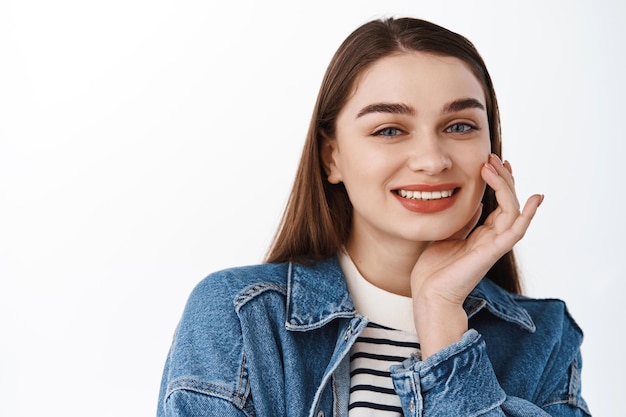 Bellezza e cura della pelle. Giovane adolescente che tocca la pelle fresca e pulita e sorride compiaciuta, tenendo la mano sulla guancia mentre guarda felice la telecamera, in piedi su sfondo bianco