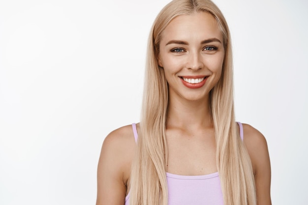 Bellezza e cura della pelle Ragazza bionda sorridente con il trucco naturale che sembra felice alla macchina fotografica in piedi in tanktop su sfondo bianco