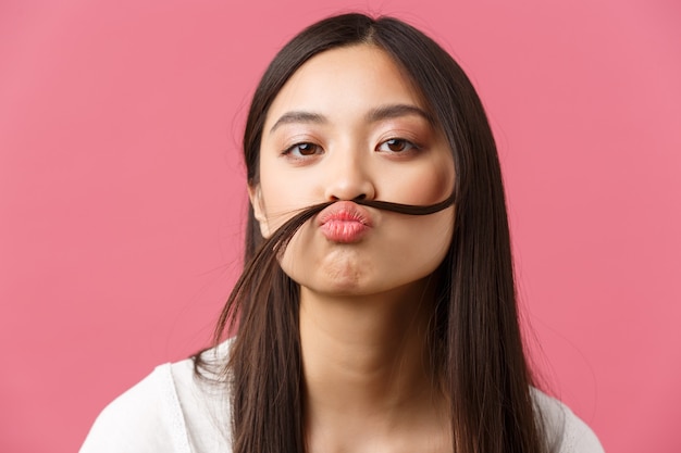 Bellezza, emozioni delle persone e concetto di svago estivo. Primo piano di una ragazza asiatica sciocca e divertente che fa baffi finti con una ciocca di capelli sul labbro, fissa la telecamera giocosa, sfondo rosa