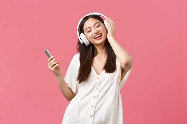 Bellezza, emozioni delle persone e concetto di tecnologia. Ragazza asiatica spensierata e carina che si gode la musica con le cuffie wireless, chiude gli occhi e sorride ascoltando la canzone preferita, tenendo in mano lo smartphone e ballando.