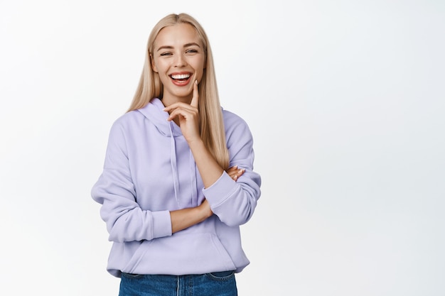 Bellezza. Giovane donna caucasica con lunghi capelli biondi, pelle pulita e sorriso bianco, che sembra felice, in piedi in felpa con cappuccio viola su bianco.