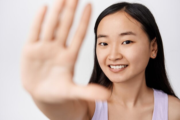 Bellezza La giovane donna asiatica sorridente con la pelle del viso sana e pulita estende il palmo mostrando il gesto di arresto della mano e guardando felice e in piedi libero su sfondo bianco