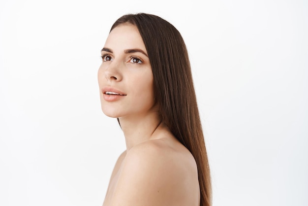 Bellezza naturale modello femminile bruna pelle pulita e lunghi capelli castani sani che guardano da parte profilo girato con pelle liscia nuda e idratata morbida lineamenti del viso con cosmetici per la cura della pelle