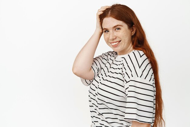 Bellezza naturale. Ragazza con i capelli rossi e le lentiggini, tocca la testa, sorride felice e guarda la telecamera, espressione del viso sicura e allegra, posa in studio bianco sullo sfondo