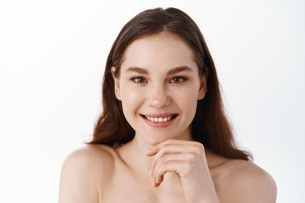 Bellezza per la cura della pelle. Giovane donna perfetta con trucco nudo naturale, spalle nude, che mostra una pelle del viso nutrita e luminosa, sorridente soddisfatto, guardandosi allo specchio dopo la doccia, sfondo bianco studio