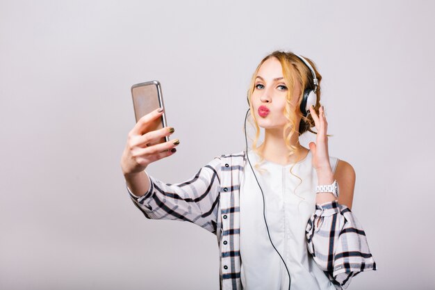 Bellissima giovane donna carina prendendo selfie vicino al muro grigio, guardando smartphone, inviando un bacio. Ragazza bionda allegra che indossa camicia a quadri e camicetta bianca. Felici, luminose emozioni. Isolato..