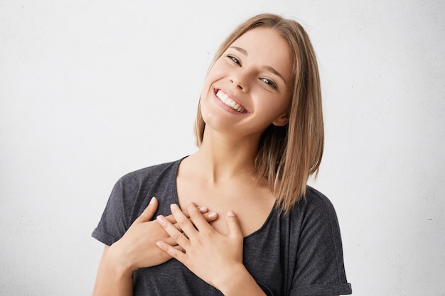 Bellissima giovane donna di razza mista dall'aspetto amichevole e positiva con un bel sorriso sincero che si sente grata e grata, mostrando il suo cuore pieno di amore e gratitudine con le mani sul seno