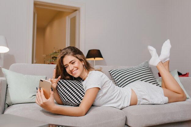 Bellissima giovane donna in calzini bianchi sdraiata sul divano e facendo selfie