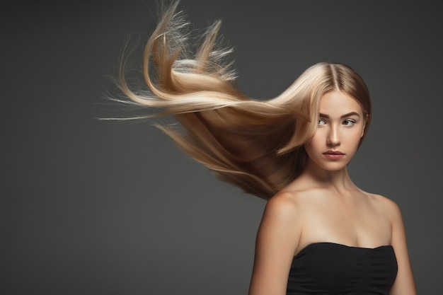 Bellissima modella con lunghi capelli biondi lisci e volanti isolati su sfondo grigio scuro dello studio. Giovane modello caucasico con pelle ben tenuta e capelli che soffia in aria.