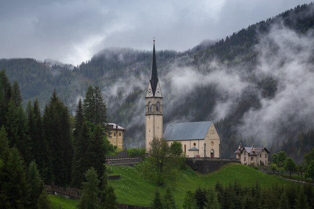 Bellissimi edifici in una foresta verde nebbiosa con alberi densi e montagne nelle Dolomiti, Italia