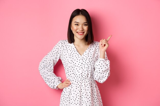 Bellissimo modello femminile asiatico sorridente, puntando il dito nell'angolo in alto a destra dello spazio della copia, mostrando banner pubblicitario, in piedi su sfondo rosa